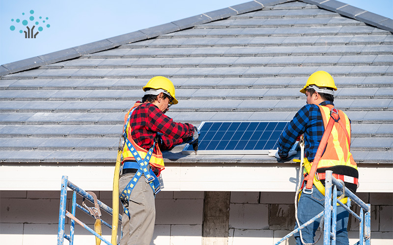 Two construction workers in safety gear installing a solar panel on a tiled roof using scaffolding.