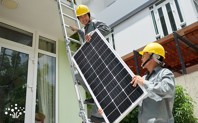 Two workers in yellow helmets installing a solar panel—one on a ladder, one handing it up—outside a modern building.