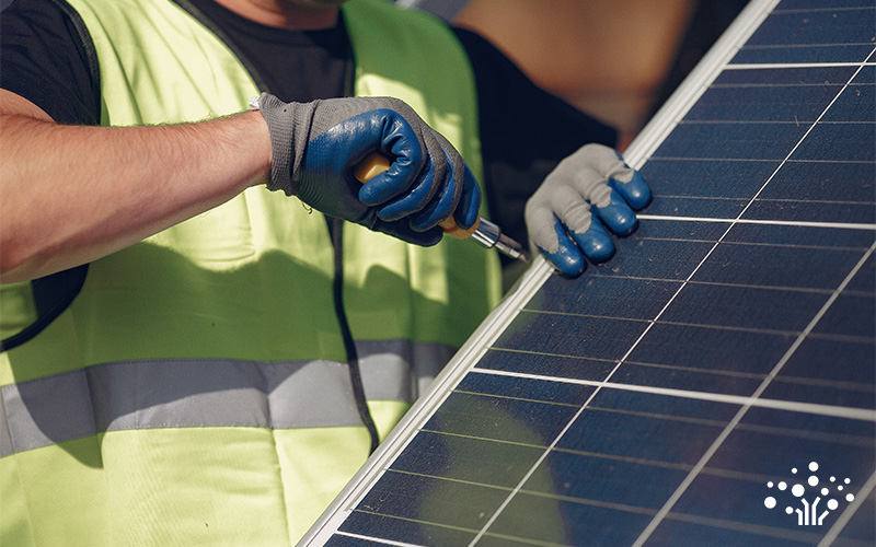 Person in safety vest and gloves using tool to work on edge of solar panel with visible photovoltaic cells.