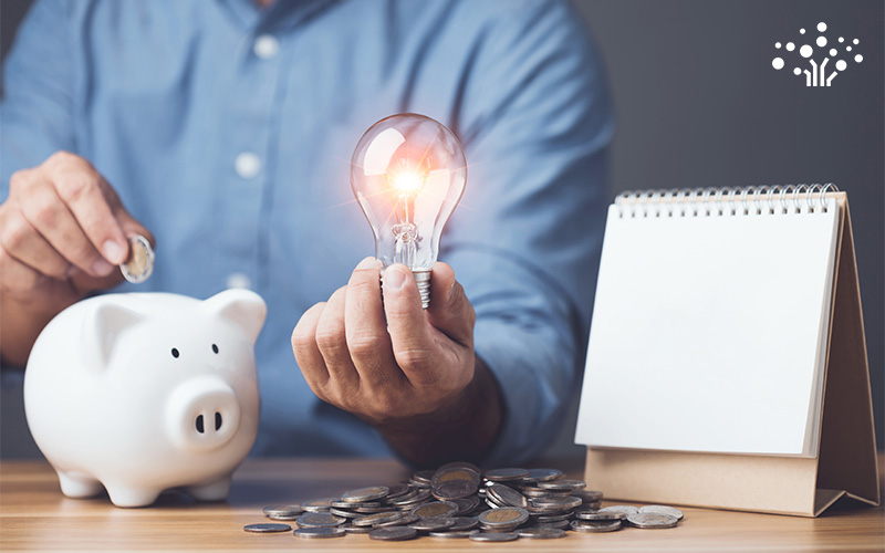 Person in blue shirt holding lit light bulb and placing coin into white piggy bank beside notepad and coins