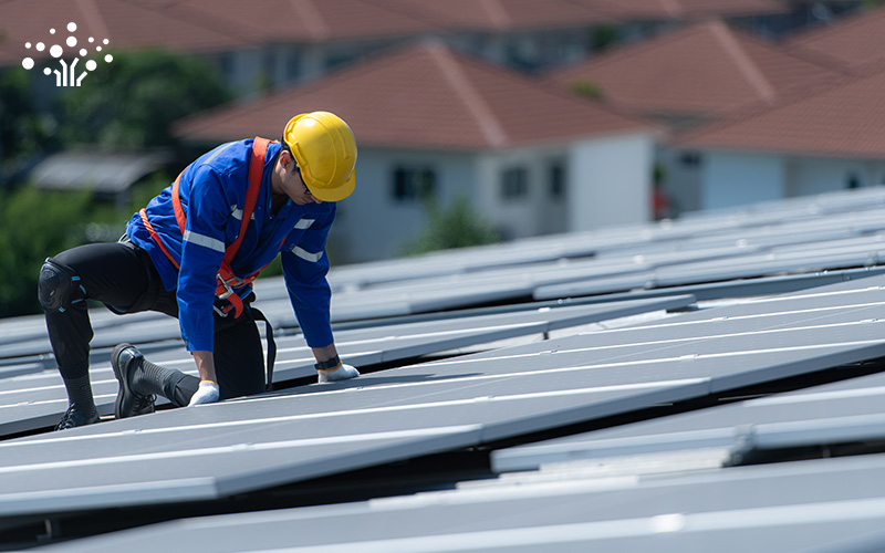 Technician with safety harness installing solar panels on warehouse roof.
